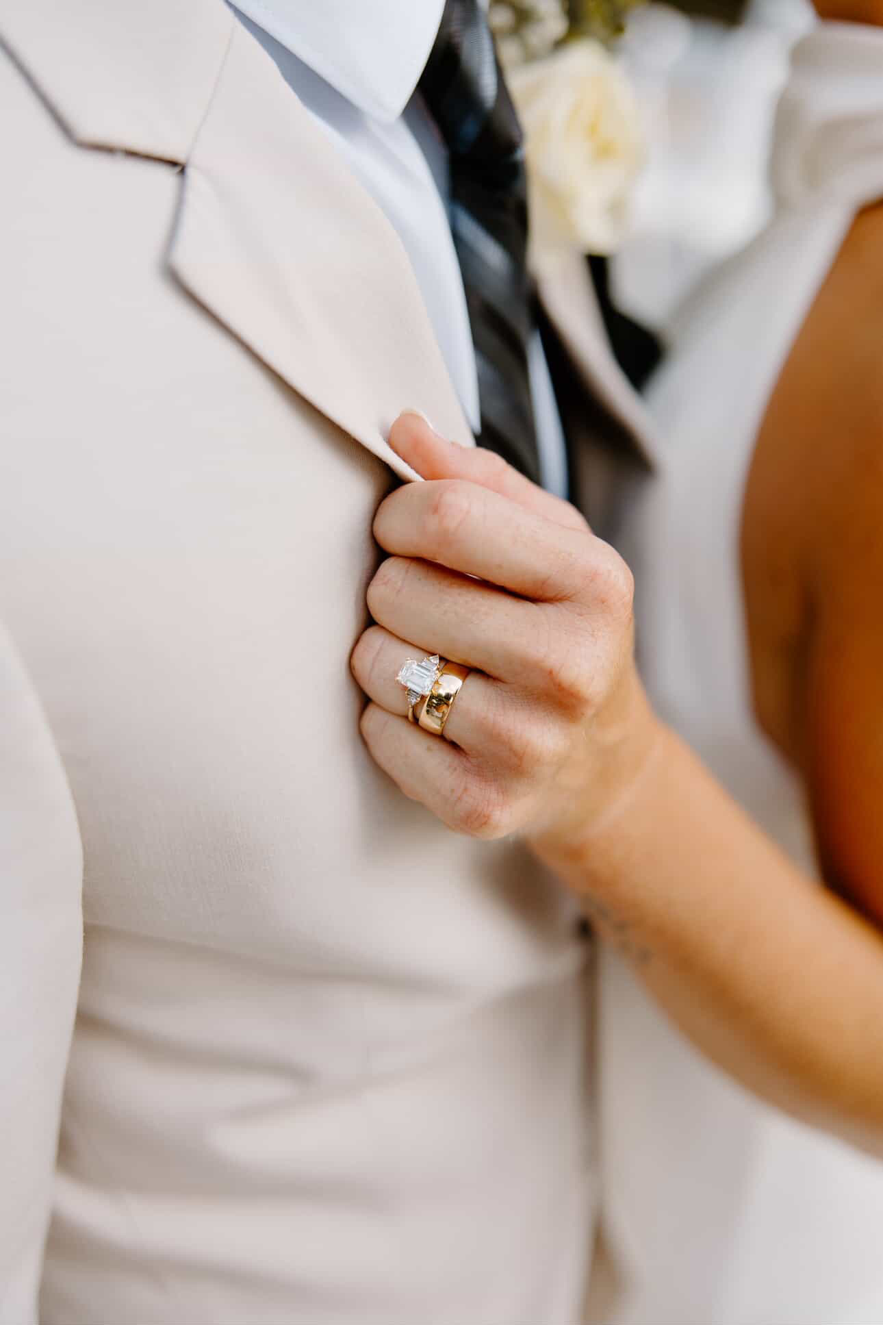 Bride's hand holding a cream suit lapel, engagement ring and wedding band visible